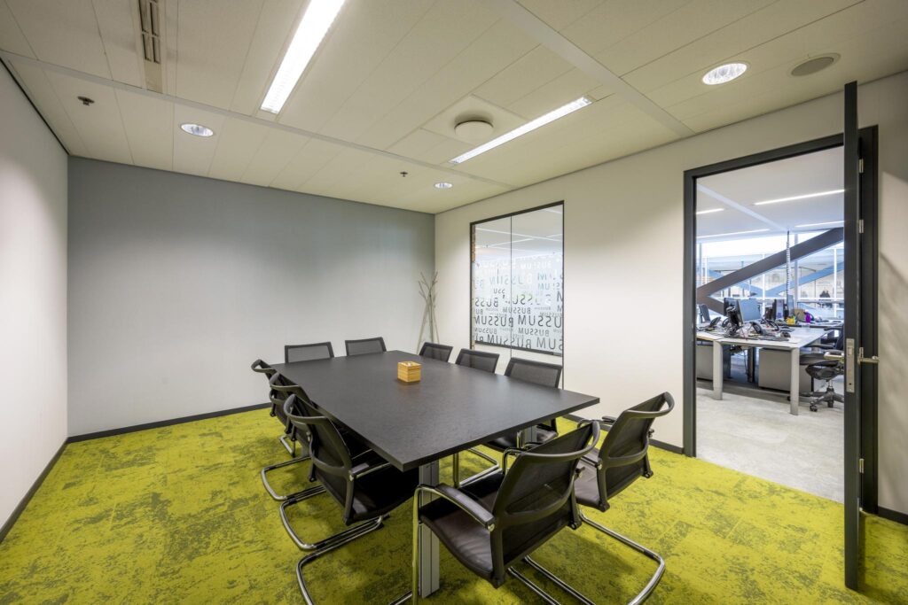 Modern conference room with a black table, eight chairs, and green carpet, connected to an open office area at Jacob Bontiusplaats.